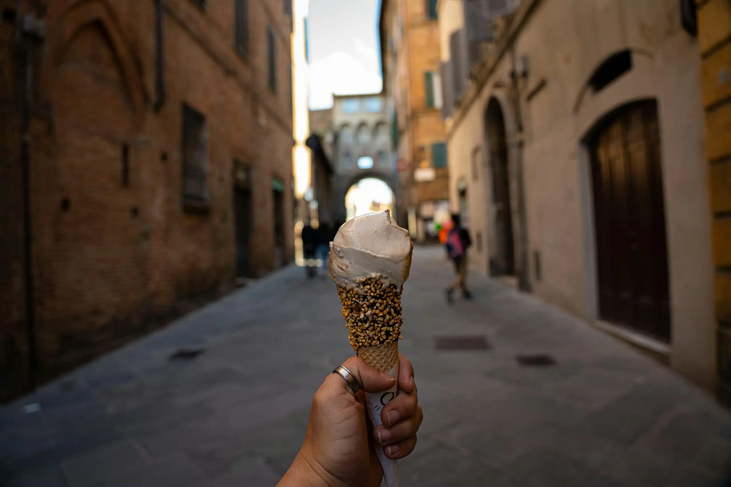 A hand holding a gelato cone rolled in chopped pistachios, with a narrow sunlit street behind