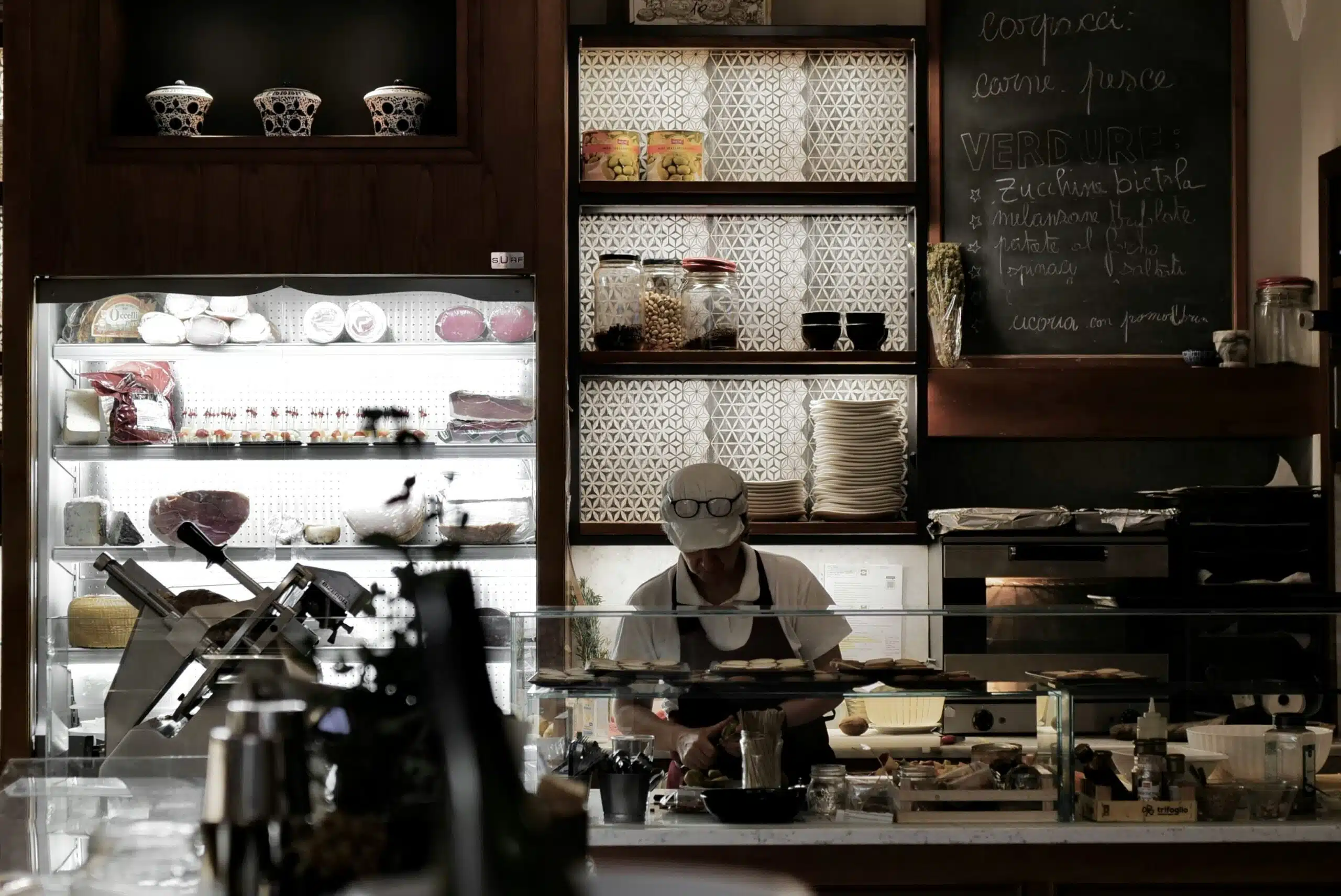 A shopkeeper in a white cap working behind the counter of a small Italian food shop lined with display cases