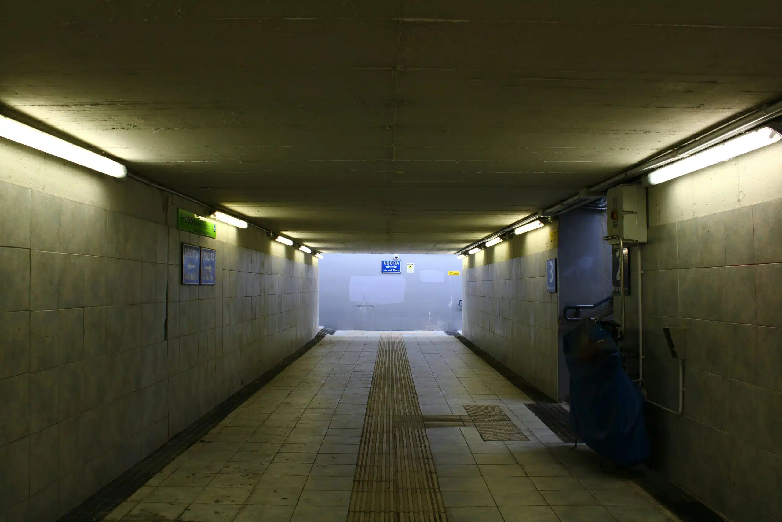 A tiled pedestrian underpass lit by fluorescent strips, leading to a bright exit at the far end