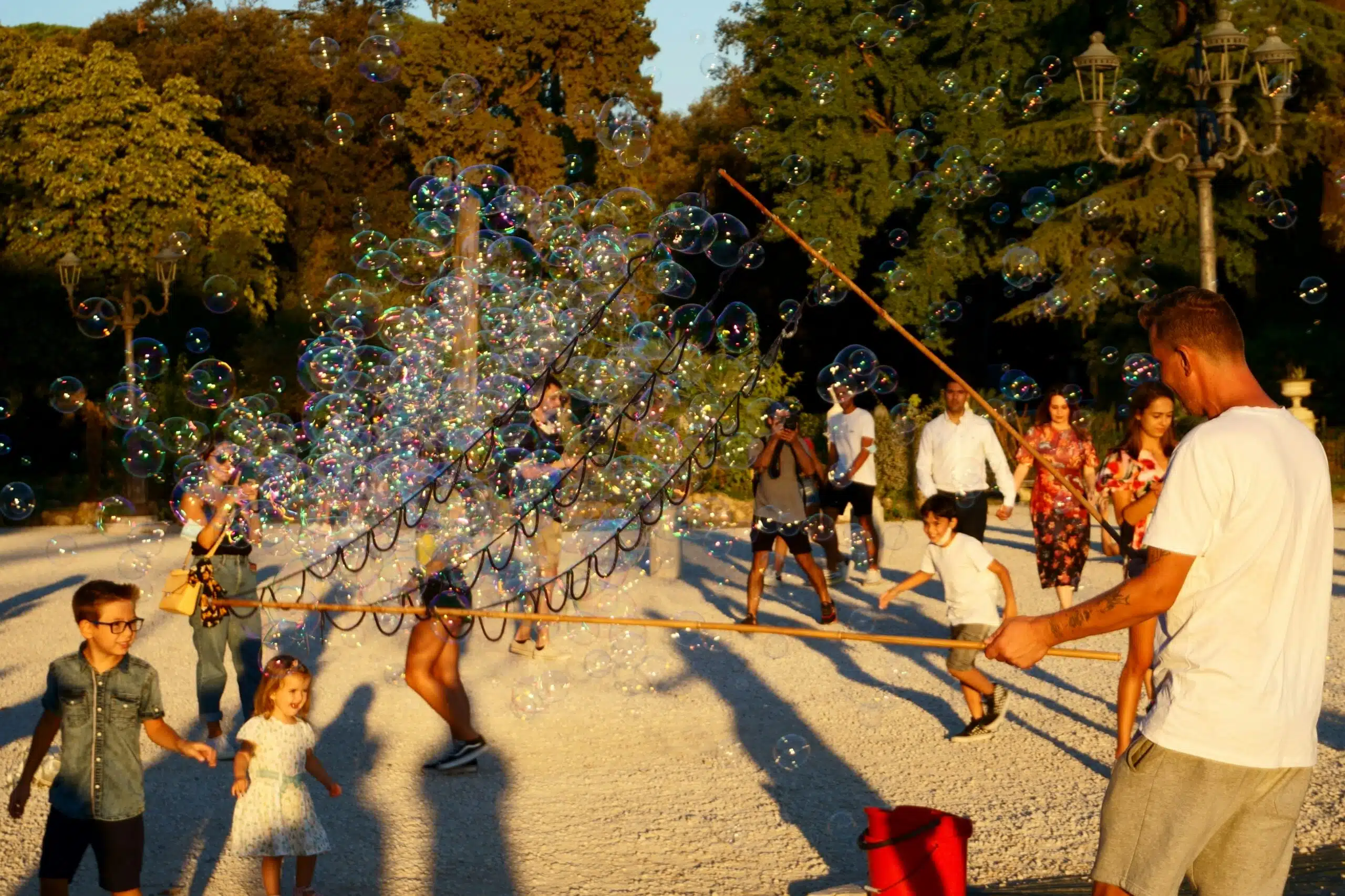 Children chasing soap bubbles in a park at golden hour while a street performer makes them with a long wand