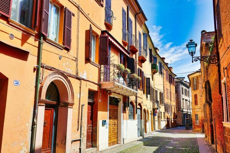 A narrow cobbled street lined with ochre and red-brick houses, shuttered windows and a wrought-iron street lamp under a blue sky