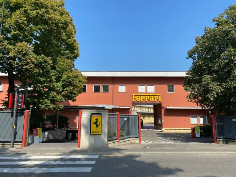 The red entrance building of the Ferrari factory in Maranello, with the yellow Ferrari sign above the gate and the prancing horse shield on the gatepost