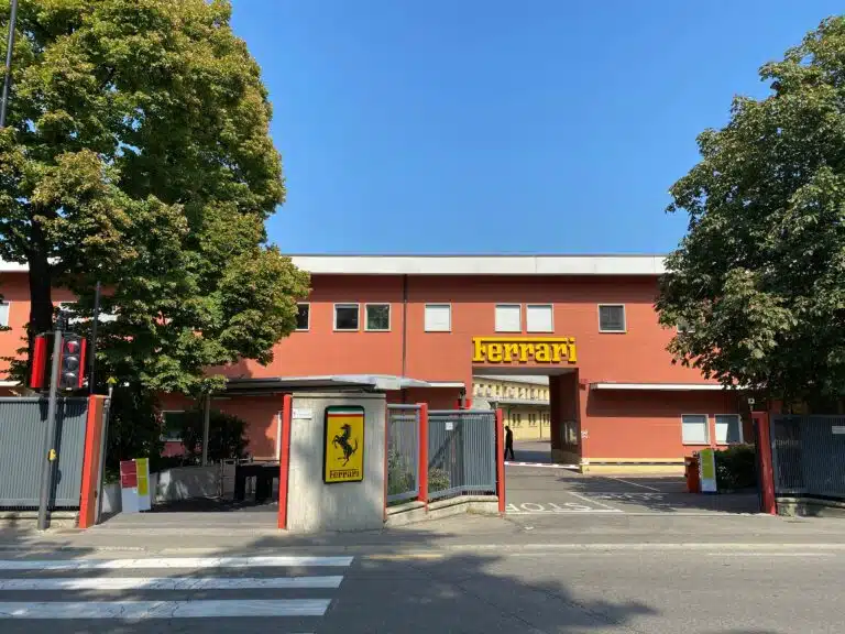 The red entrance building of the Ferrari factory in Maranello, with the yellow Ferrari sign above the gate and the prancing horse shield on the gatepost