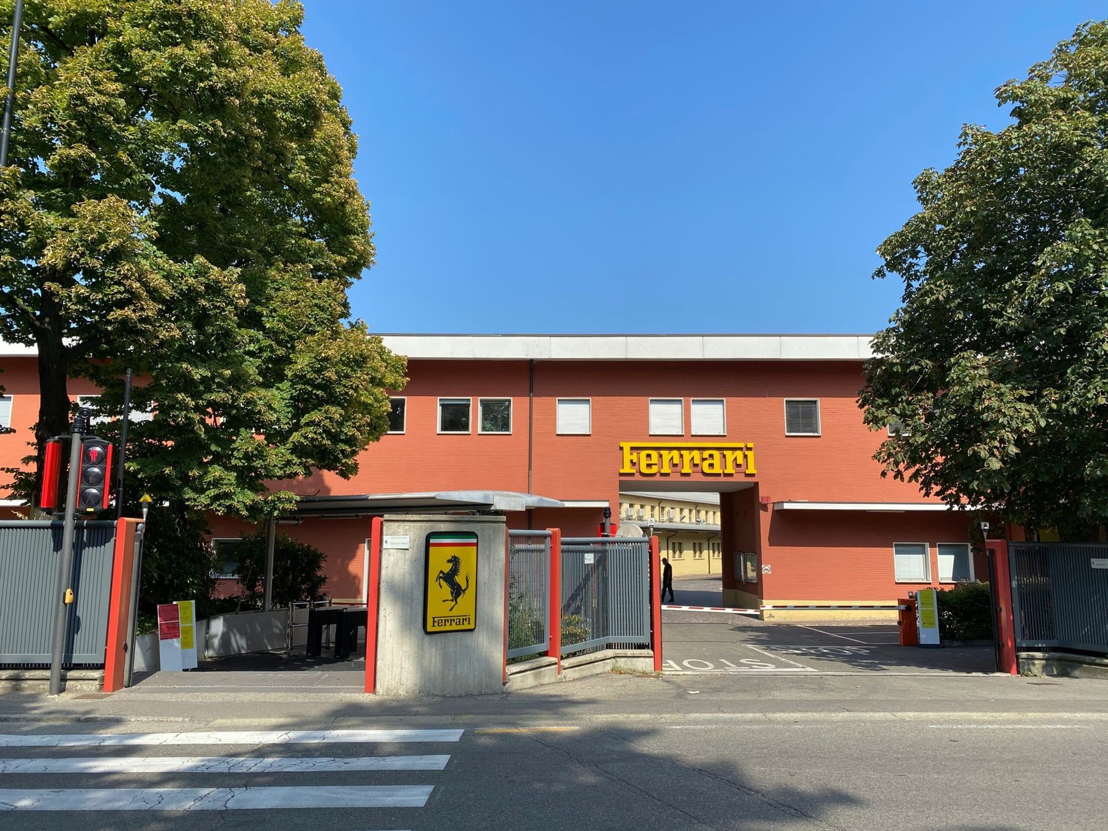 The red entrance building of the Ferrari factory in Maranello, with the yellow Ferrari sign above the gate and the prancing horse shield on the gatepost