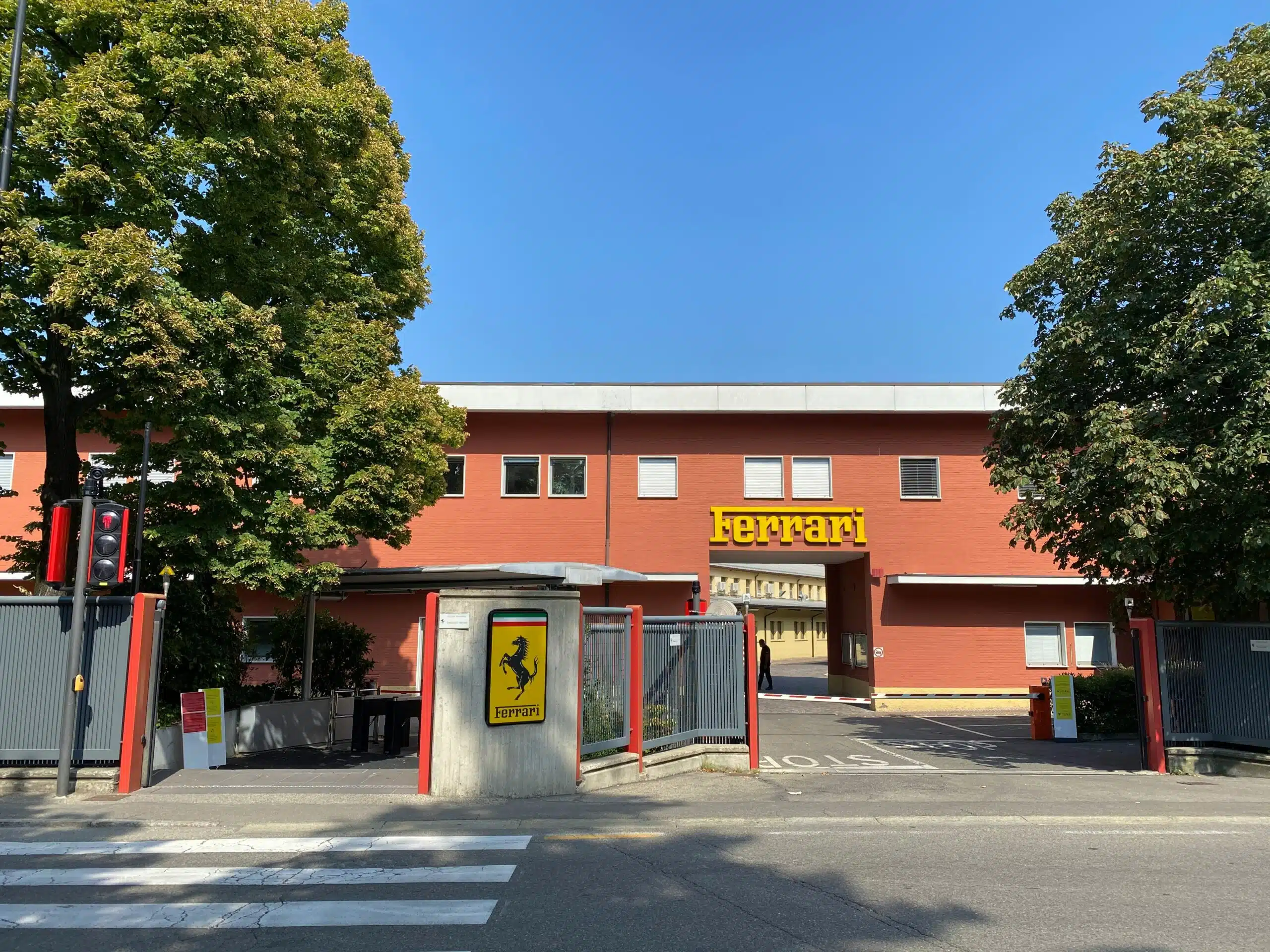 The red entrance building of the Ferrari factory in Maranello, with the yellow Ferrari sign above the gate and the prancing horse shield on the gatepost